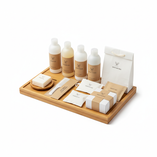 Complete hotel bathroom amenities set on bamboo tray overhead view showing all items: shampoo, shower gel, conditioner, lotion, soap, dental kit, comb, shower cap, shaving kit, vanity kit, cotton pads and sanitary bag in matching white and kraft packaging