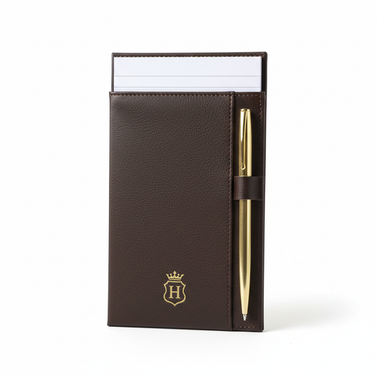 Dark brown PU leather hotel notepad holder standing upright with white hotel notepad inserted at top and gold ballpoint pen in side pen slot, subtle gold embossed hotel logo on front face on white background, compact hospitality guestroom desk stationery organizer
