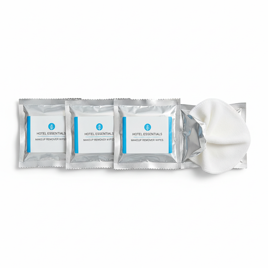 Hotel makeup remover wipes in individual foil sachet packets, one open showing soft nonwoven wipe, three sachets arranged on white background, guest bathroom personal care amenity