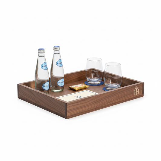 Dark walnut wood hotel mini bar tray with low side rails holding two mineral water bottles two drinking glasses wrapped snack package and folded hotel information card with laser-engraved logo on end panel on white background, custom hospitality guestroom minibar organizer