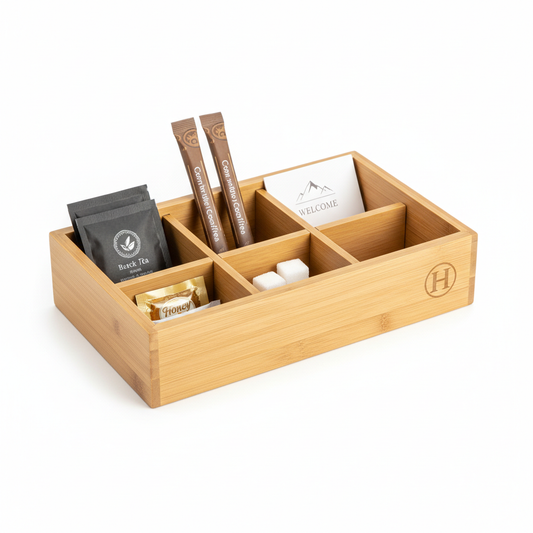 Natural light bamboo hotel amenity box open-top tray with multiple compartments neatly holding tea sachets coffee sticks honey packet sugar cubes and small card with laser-engraved hotel logo on end panel on white background, custom hospitality guestroom supply organizer